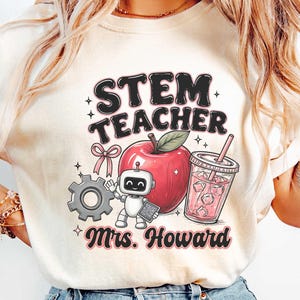 May include: An ivory t-shirt featuring a graphic with the text STEM TEACHER and Mrs. Howard. The design includes a red apple, a small grey robot, a grey gear, a pink bow, and a clear cup with a pink straw filled with ice cubes and pink liquid.