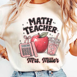 May include: An ivory t-shirt featuring a graphic with the text MATH TEACHER and Mrs. Miller. The design includes a pink calculator with a bow, a red apple with a green leaf, a ruler, and a pink drink in a clear cup with a straw and ice cubes.