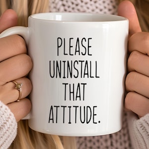 May include: White ceramic mug with the text "PLEASE UNINSTALL THAT ATTITUDE" in black. The mug is held by a person wearing a cream-colored sweater. The mug has a standard handle.
