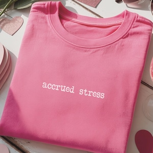 May include: A bright pink t-shirt with the words "accrued stress" in white lettering. The shirt is folded neatly on a white wooden surface, surrounded by heart-shaped decorations, sunglasses, and a glass of pink liquid.