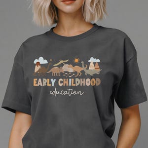 May include: Dark gray t-shirt with a graphic featuring dinosaurs, clouds, and a sun. The text "EARLY CHILDHOOD education" is printed in a brown and gold color scheme. The shirt is a casual, short-sleeved design.