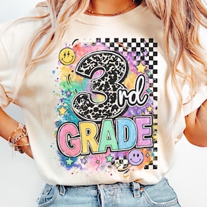 May include: An ivory t-shirt featuring a graphic print with the text 3rd GRADE. The design includes a leopard print number 3, colorful watercolor splashes, a black and white checkered pattern, yellow stars, and two yellow and purple smiley faces.