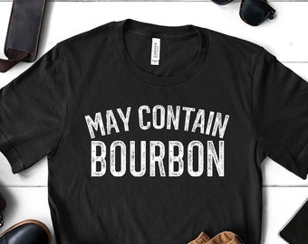 May include: Black t-shirt with white text that reads "May Contain Bourbon". The shirt is laid flat on a white wooden surface with brown pants, a pair of sunglasses, and a key chain.