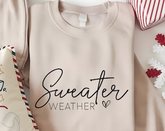 May include: A sand-colored sweatshirt with the words "Sweater Weather" in black script. A heart symbol follows the word "Weather". The sweatshirt is next to a decorative pillow and a wrapped gift.