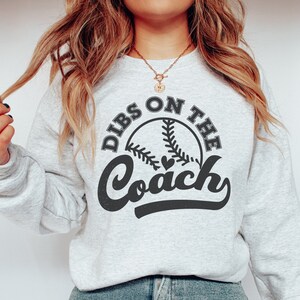 May include: A gray sweatshirt with the text "Dibs on the Coach" in black lettering, featuring a baseball graphic with a heart.