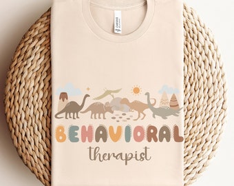May include: A beige t-shirt with a design of dinosaurs and the text "BEHAVIORAL therapist" in colorful letters.