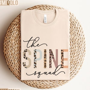 May include: A beige t-shirt with the text "the SPINE squad" in black script. The letters in "SPINE" are decorated with a leopard print pattern in various colors.