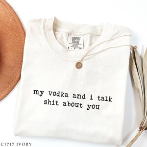May include: Cream-coloured Comfort Colors t-shirt with the text "my vodka and i talk shit about you" in black. A gold necklace with a pendant is draped across the shirt. A brown hat is visible in the background.