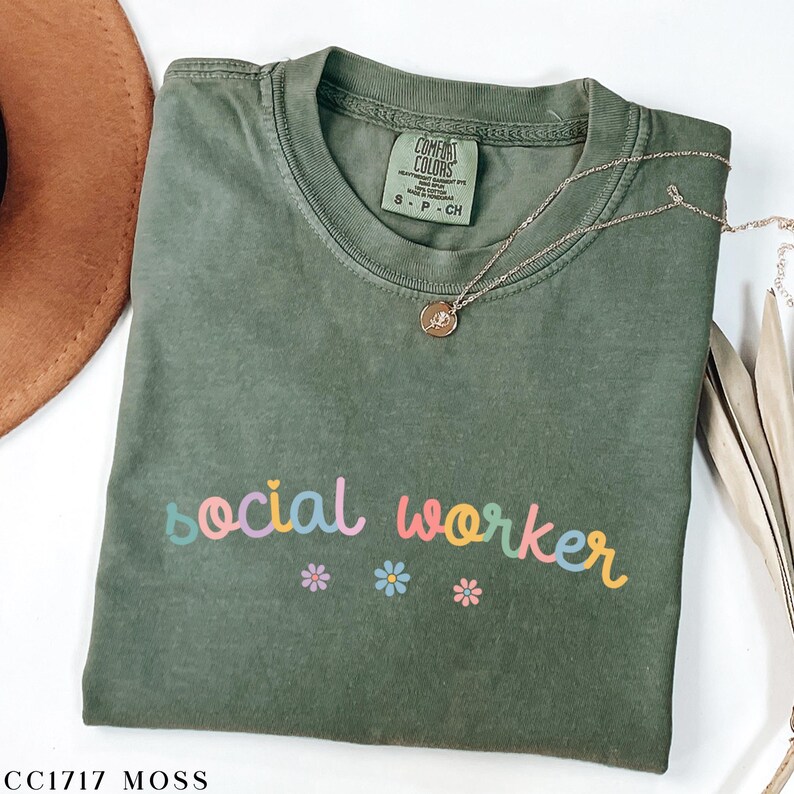 Pode incluir: Uma camiseta verde musgo com as palavras "social worker" em cores do arco-&iacute;ris. A camiseta tem um pequeno desenho de flores abaixo do texto.