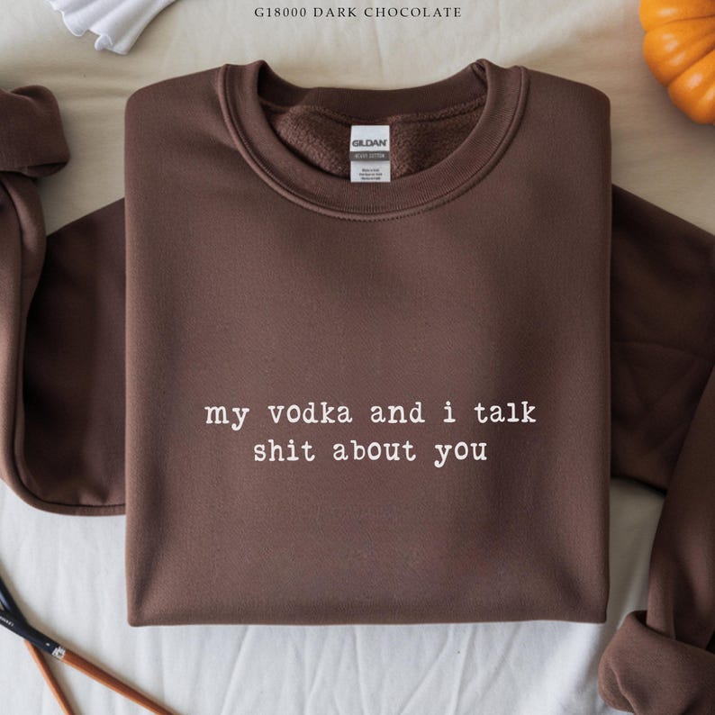 May include: A dark chocolate-coloured sweatshirt with the text "my vodka and i talk shit about you" in white. The Gildan brand label is visible on the upper centre of the sweatshirt. The sweatshirt is on a white surface.