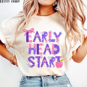 May include: Ivory-colored t-shirt with the text "EARLY HEAD START" in purple and pink block letters. The design includes floral and polka dot patterns, a pink bow, and a pink apple graphic. The shirt is a casual, everyday wear item.