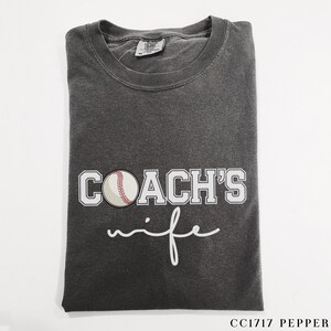 Może przedstawiać: Szary t-shirt z napisem "COACH'S wife" w białym kolorze. Biała piłka baseballowa z czerwonymi szwami znajduje się nad tekstem.