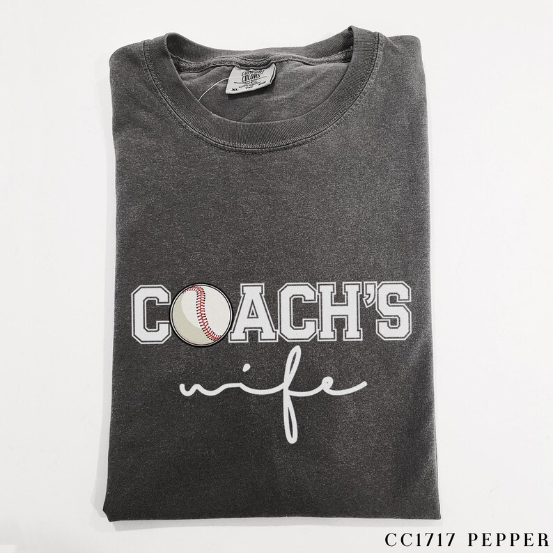 Może przedstawiać: Szary t-shirt z napisem "COACH'S wife" w białym kolorze. Biała piłka baseballowa z czerwonymi szwami znajduje się nad tekstem.