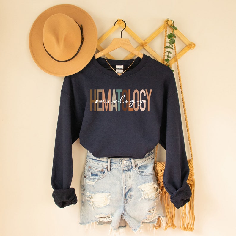 May include: Navy blue sweatshirt with the text "HEMATOLOGY oncology" in colorful letters. The sweatshirt is hanging on a wooden hanger with a straw hat and a woven bag hanging nearby.