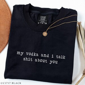 May include: Black t-shirt with the text "my vodka and i talk shit about you" in white. A gold-coloured necklace with a round pendant rests on the shirt. The shirt has a crew neck and short sleeves. The brand label is visible.