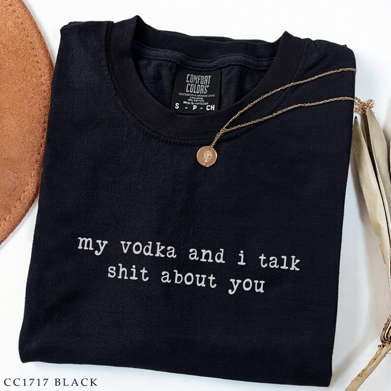 May include: Black t-shirt with the text "my vodka and i talk shit about you" in white. A gold-coloured necklace with a round pendant rests on the shirt. The shirt has a crew neck and short sleeves. The brand label is visible.