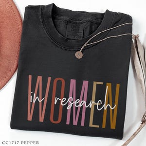 May include: A black t-shirt with the words "WOMEN in research" printed in a colorful design. A silver necklace with a pendant rests on the shirt. The shirt is labeled "CC1717 PEPPER".