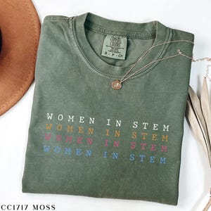 May include: An olive green t-shirt with the text "WOMEN IN STEM" in a colorful, stacked design. The shirt has a crew neck and short sleeves. A gold necklace with a pendant rests on the shirt.
