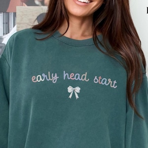 May include: A teal green sweatshirt with the text "early head start" in rainbow colors and a white bow with pink and purple polka dots.