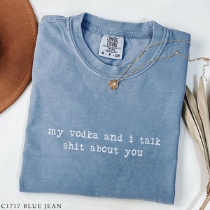 May include: A blue jean Comfort Colors t-shirt with the text "my vodka and i talk shit about you" in white. A gold necklace with a pendant is draped across the shirt. The shirt is folded and has a small tag.