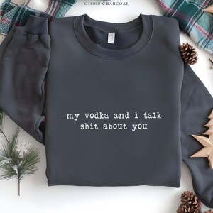 May include: Dark grey sweatshirt featuring the white text "my vodka and i talk shit about you". The top has a crew neck and long sleeves. Festive decorations are visible in the background.