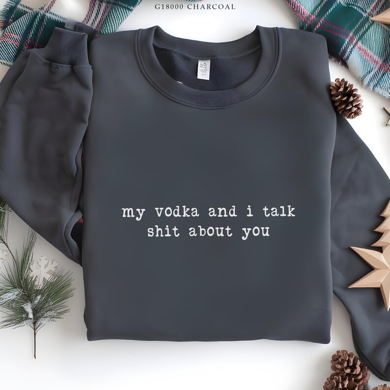 May include: Dark grey sweatshirt featuring the white text "my vodka and i talk shit about you". The top has a crew neck and long sleeves. Festive decorations are visible in the background.