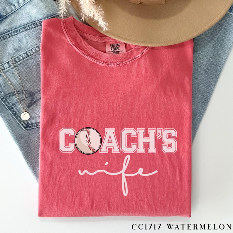 Może przedstawiać: Czerwony T-shirt z napisem "COACH'S wife" w kolorze białym i grafiką r&oacute;żowej piłki baseballowej. Koszulka jest wykonana z wygodnej mieszanki bawełny.