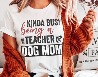 Puede incluir: Camiseta blanca con el texto "KINDA BUSY being a TEACHER & DOG MOM" en letras negras y burdeos. La persona lleva una rebeca color óxido. Una taza blanca está en primer plano.