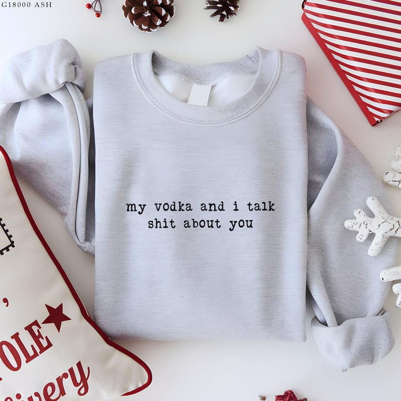 May include: A pale grey sweatshirt with the text "my vodka and i talk shit about you" in black. The sweatshirt is surrounded by festive decorations, including pine cones and a gift box.