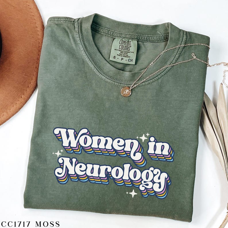 May include: A green t-shirt with the text "Women in Neurology" in a retro rainbow font. The shirt has a distressed look and is made of a soft, comfortable fabric.