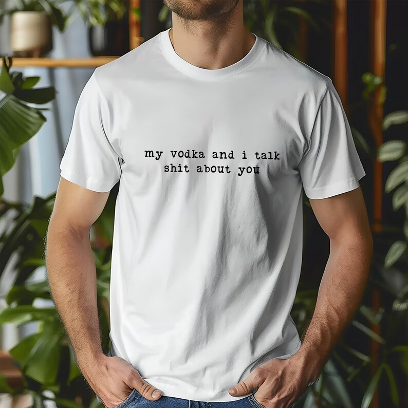 May include: White t-shirt with the text "my vodka and I talk shit about you" in black, sans-serif font. The shirt is short-sleeved and appears to be made of cotton. The person is wearing blue jeans.