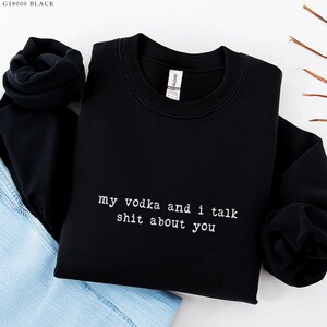 May include: Black sweatshirt with the text "my vodka and i talk shit about you" in white. The sweatshirt is folded with a pair of blue jeans.