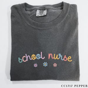May include: A dark gray t-shirt with the text "school nurse" in colorful letters and three small flower designs below the text.