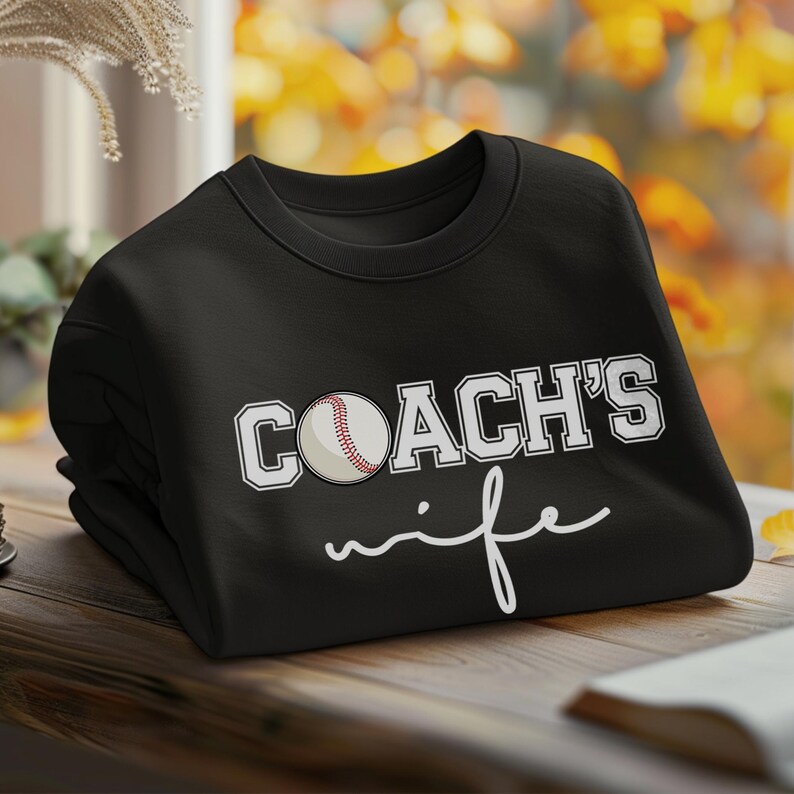 Może przedstawiać: Czarna bluza z białym napisem "COACH'S wife" i grafiką baseballa.