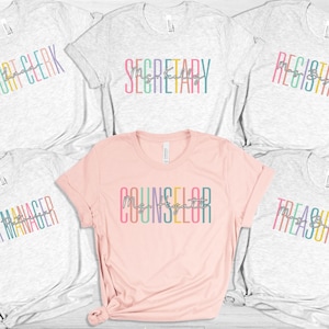 May include: Set of six grey and pink t-shirts with rainbow text that reads "Court Clerk", "Secretary", "Registrar", "Data Manager", "Counselor", and "Treasurer".