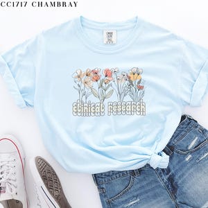 May include: Light blue t-shirt with a floral design and the text "clinical research" in a retro font.
