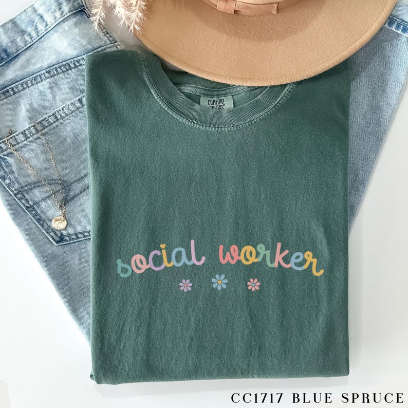Pode incluir: Uma camiseta verde com o texto "social worker" em cores do arco-&iacute;ris com pequenos detalhes de flores. A camiseta &eacute; feita de um material macio e confort&aacute;vel.