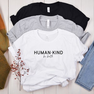May include: White t-shirt with black text that reads "HUMAN:KIND be both". The shirt is folded and displayed on a white wooden surface with a sprig of red berries and a pair of blue jeans.