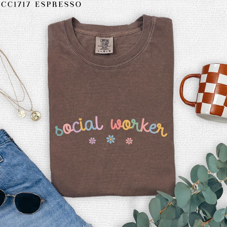 Pode incluir: Uma camiseta marrom com o texto "social worker" em cores do arco-&iacute;ris e tr&ecirc;s pequenas flores rosa, azuis e amarelas.
