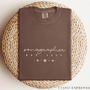 May include: A brown t-shirt with the word "sonographer" in a cursive font, along with "EST. 2025" and small flower graphics. The shirt is displayed on a woven, circular surface. The shirt's color is "CC1717 Espresso".