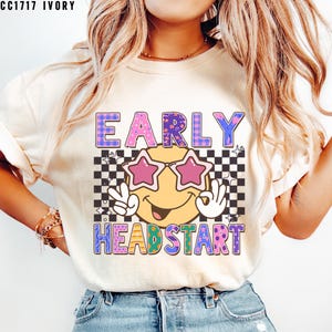 May include: Ivory-colored t-shirt with the text "EARLY HEADSTART" in a colorful, retro design. The graphic includes a smiley face wearing star-shaped sunglasses and making peace signs, set against a checkered background.