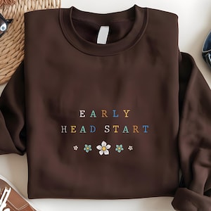May include: A brown sweatshirt with the words "EARLY HEAD START" in colorful letters. Below the text are small flower graphics. The sweatshirt is folded and appears to be made of a soft material.