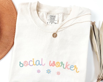 Könnte beinhalten: Ein cremefarbenes T-Shirt mit dem Text "social worker" in Regenbogenfarben und kleinen Blumenakzenten. Das Shirt ist gefaltet und liegt flach auf einer weißen Oberfläche.