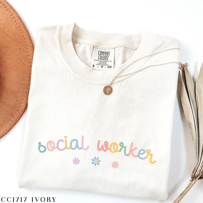 Pode incluir: Camiseta de cor marfim com o texto "social worker" em cores arco-&iacute;ris e pequenos detalhes de flores. A camiseta est&aacute; dobrada e colocada sobre uma superf&iacute;cie branca.