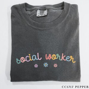 Pode incluir: Uma camiseta cinza escura com o texto "social worker" em cores do arco-&iacute;ris. O texto est&aacute; em uma fonte de escrita manuscrita divertida. Tr&ecirc;s pequenas flores rosa, amarelas e azuis est&atilde;o abaixo do texto.