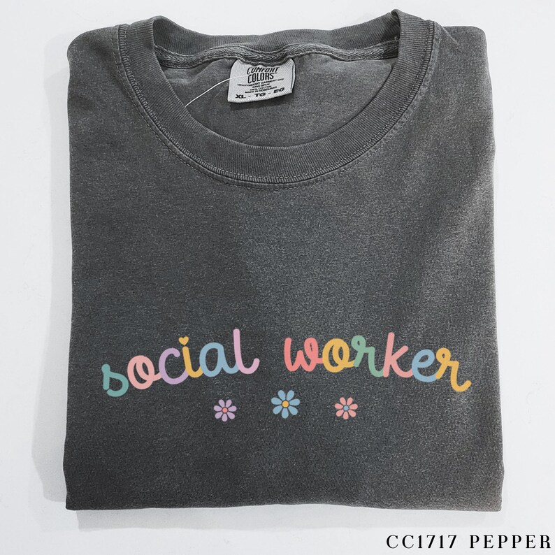 Pode incluir: Uma camiseta cinza escura com o texto "social worker" em cores do arco-&iacute;ris. O texto est&aacute; em uma fonte de escrita manuscrita divertida. Tr&ecirc;s pequenas flores rosa, amarelas e azuis est&atilde;o abaixo do texto.