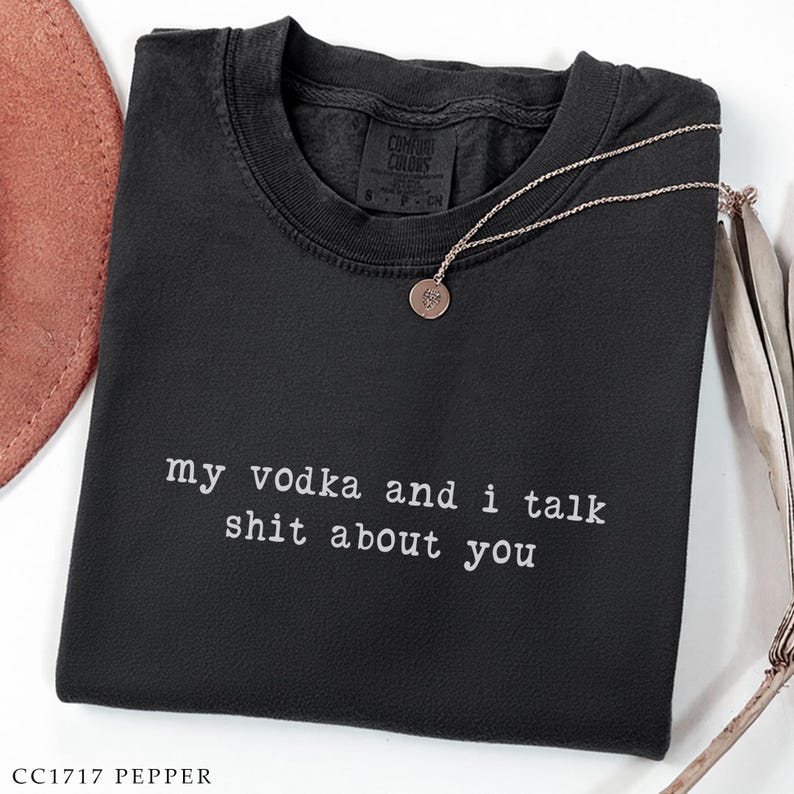 May include: Dark grey crew neck sweatshirt featuring the white text "my vodka and i talk shit about you". A silver necklace with a circular pendant rests on the shirt. The label reads "Comfort Colors".