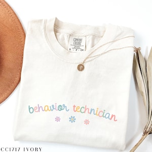 May include: Ivory t-shirt with the text "behavior technician" in rainbow colors and three small flower graphics. The shirt is folded and has a tag that says "Comfort Colors".