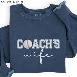Może przedstawiać: Niebieska bluza z napisem "COACH'S wife" w białym kolorze. Biała piłka baseballowa z czerwonymi szwami znajduje się na środku nad tekstem.