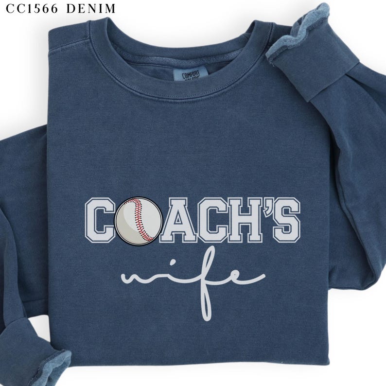 Może przedstawiać: Niebieska bluza z napisem "COACH'S wife" w białym kolorze. Biała piłka baseballowa z czerwonymi szwami znajduje się na środku nad tekstem.